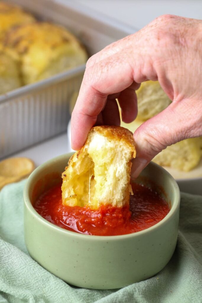 Hand dipping a sourdough cheese and garlic roll into a bowl of marinara sauce.