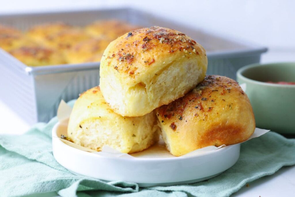 Three sourdough cheese and garlic rolls on a small plate with a pan of rolls in the background.