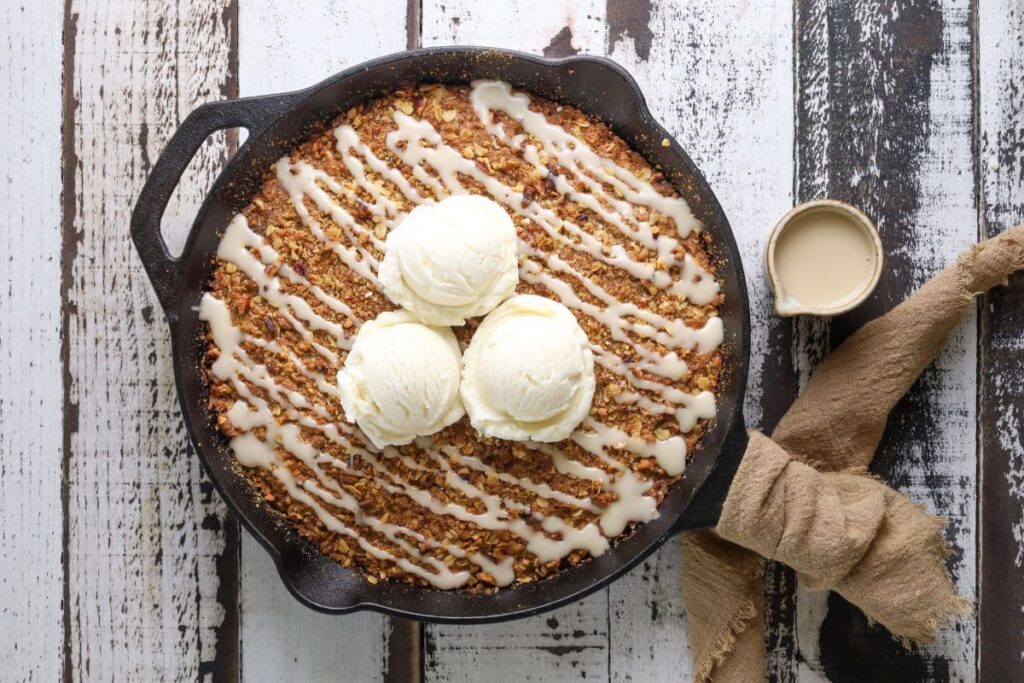 Sourdough oatmeal pumpkin cookie skillet on a wooden table with 3 vanilla ice creams scoops on top and a jar of maple glaze adjacent.