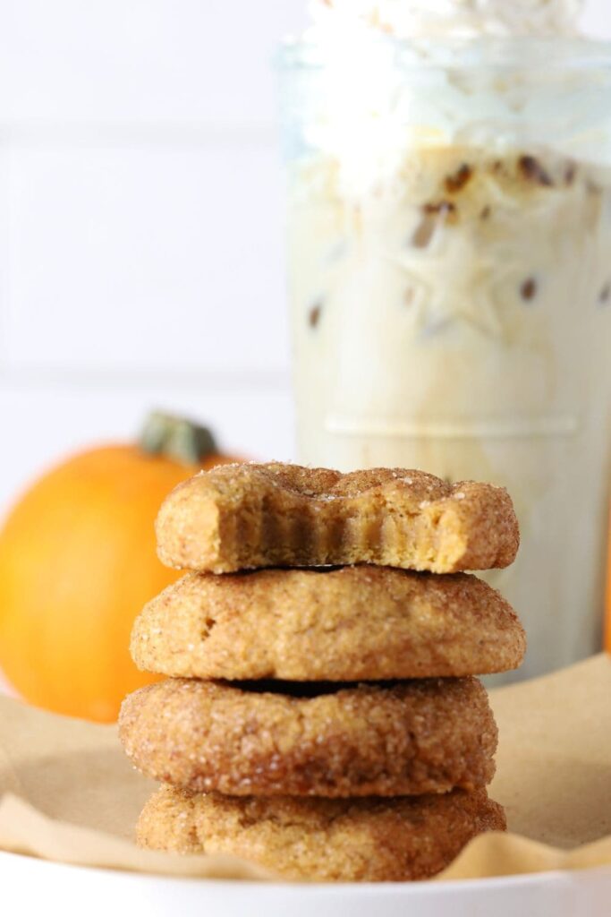 Four sourdough pumpkin snickerdoodle cookies stacked on parchment paper with a coffee drink in background.