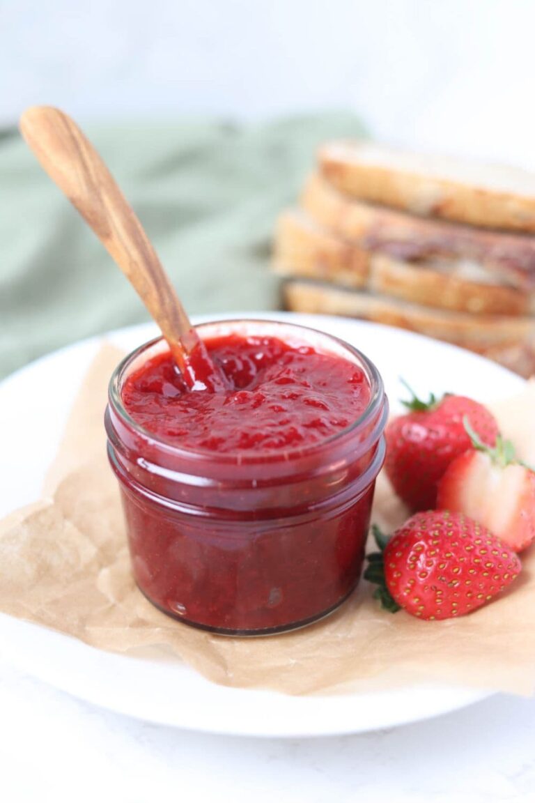 A small jar of strawberry jam sitting on a white plate alongside some fresh strawberries. There is a wooden spoon in the jar and some sourdough in the background.