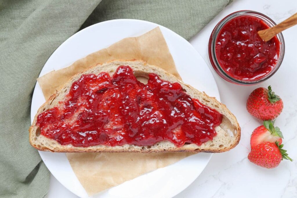 A slice of sourdough bread spread with strawberry jam sitting on a white plate.