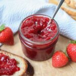 A small jar of strawberry jam with a spoon in it. There are fresh strawberries scattered around it.