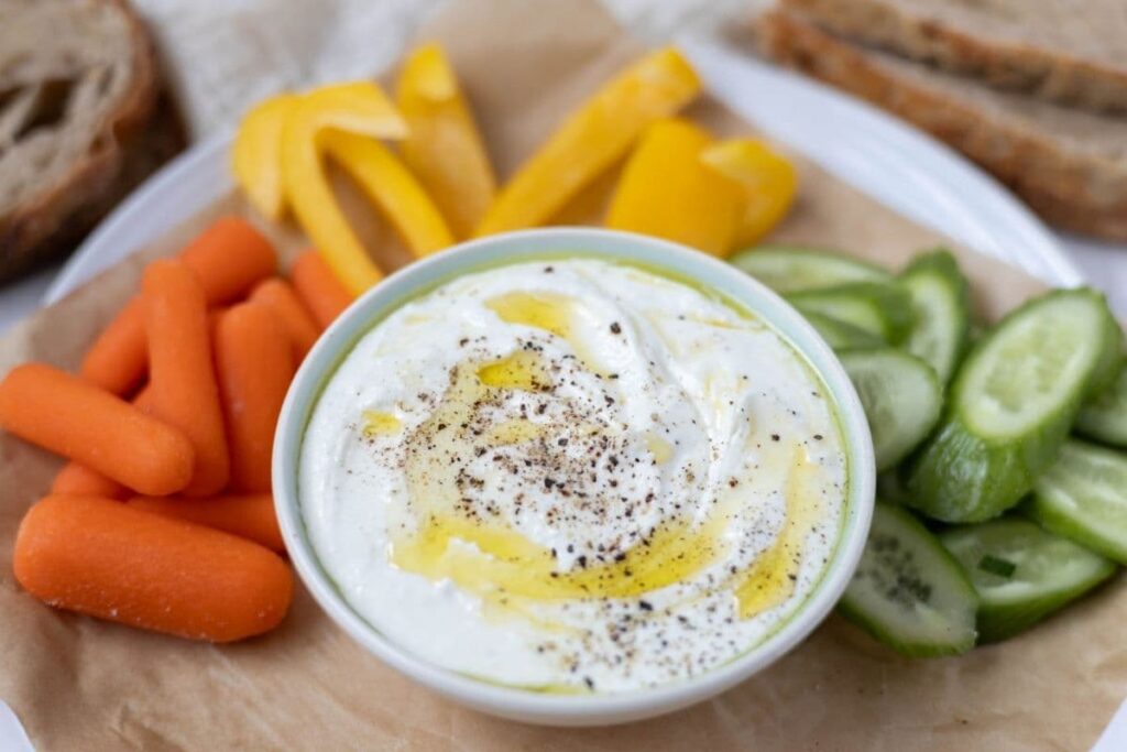 A bowl filled with whipped feta dip topped with olive oil and a spinkle of black pepper. The bowl of whipped feta dip is surrounded by carrots, yellow peppers and cucumber slices.