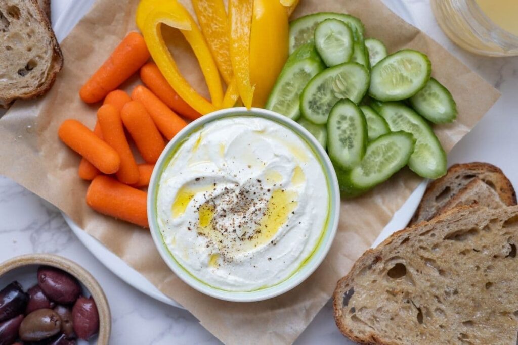 A bowl of whipped feta dip topped with olive oil and black pepper. The bowl has been served with carrots, yellow peppers, cucumber slices, black olives and whole wheat sourdough bread slices.