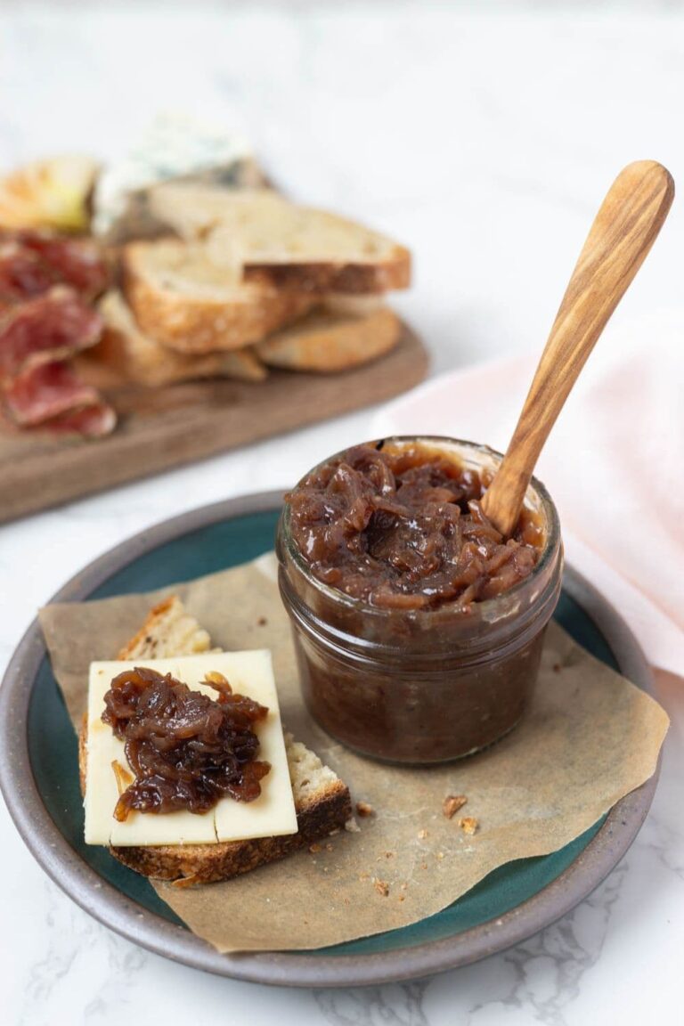 Jar of caramelized onion relish on a parchment paper lined plate next to a piece of sourdough bread topped with cheese and onion relish on top.