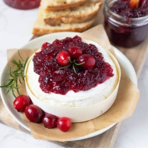 Bright red cranberry vanilla jam sitting on top of a wheel of Brie decorated with fresh cranberries and rosemary.